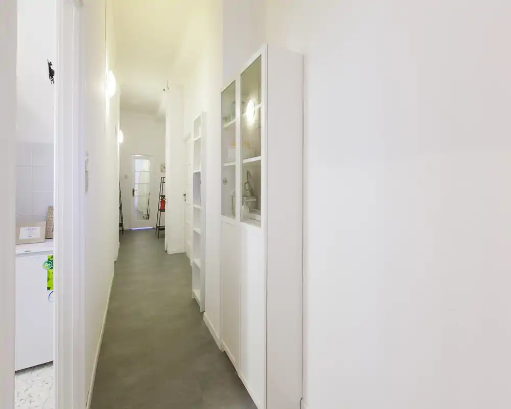 Ingresso/corridoio stretto e ben illuminato con armadietti a muro e una stanza laterale piastrellata visibile; pareti bianche e pavimento grigio per un aspetto pulito e minimale.