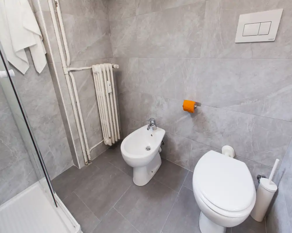 Bagno moderno e pulito con WC, bidet, termosifone e doccia visibili; piastrelle grigie neutre e disposizione compatta offrono uno spazio pratico e ben illuminato.