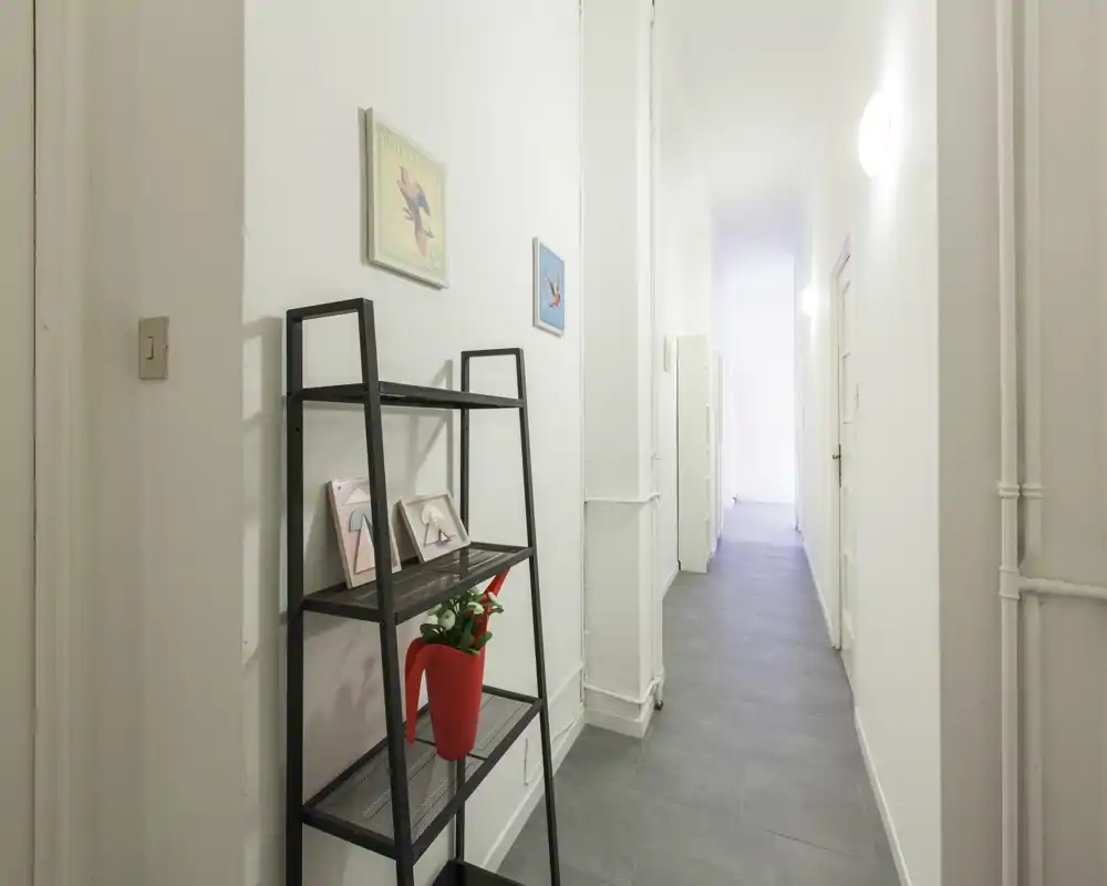 Corridoio d'ingresso stretto e luminoso con pavimento piastrellato, pareti bianche e una scaffalatura decorativa con cornici e un vaso rosso.