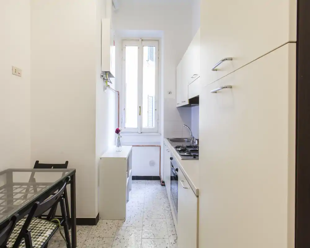 Cucina compatta a corridoio con mobili bianchi, piano cottura a gas, forno incassato e una finestra che garantisce luce naturale. Piccolo tavolo da pranzo e arredi essenziali rendono l'ambiente funzionale e luminoso.