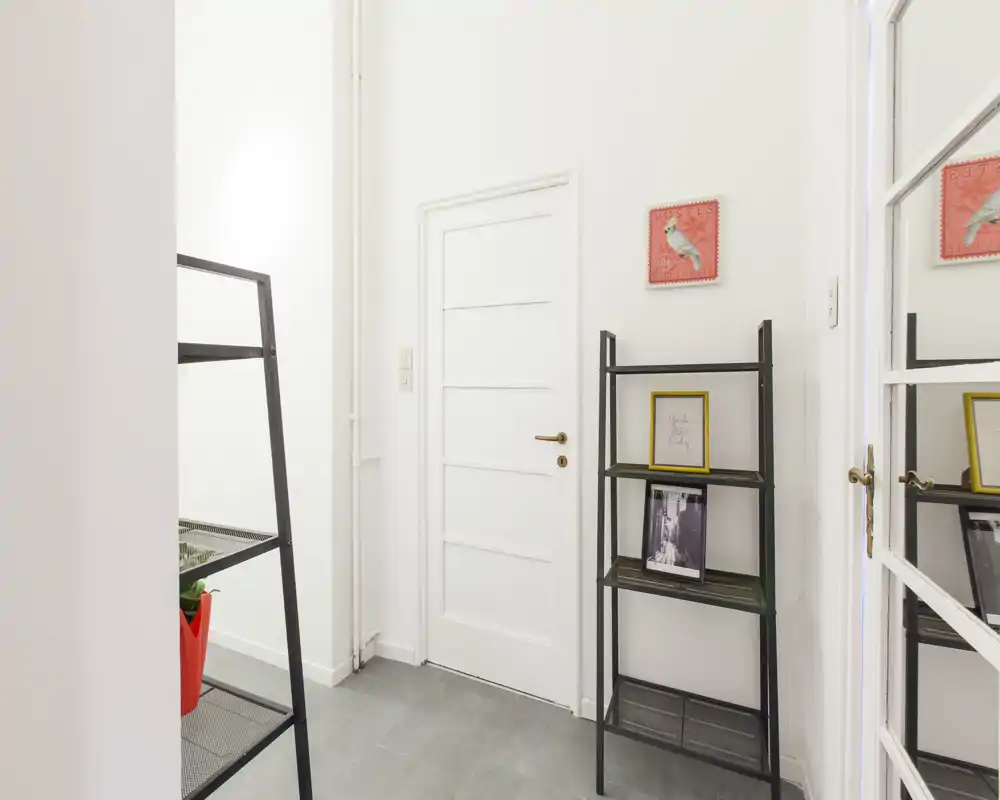 Ingresso luminoso e minimal con porta bianca, pavimento piastrellato e scaffalatura sottile per oggetti decorativi.