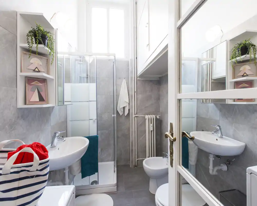 Bagno moderno e luminoso con cabina doccia, lavabo a muro, bidet e piastrelle grigie neutre. Specchio ampio, portasciugamani e mensole decorative rendono lo spazio funzionale e ordinato.