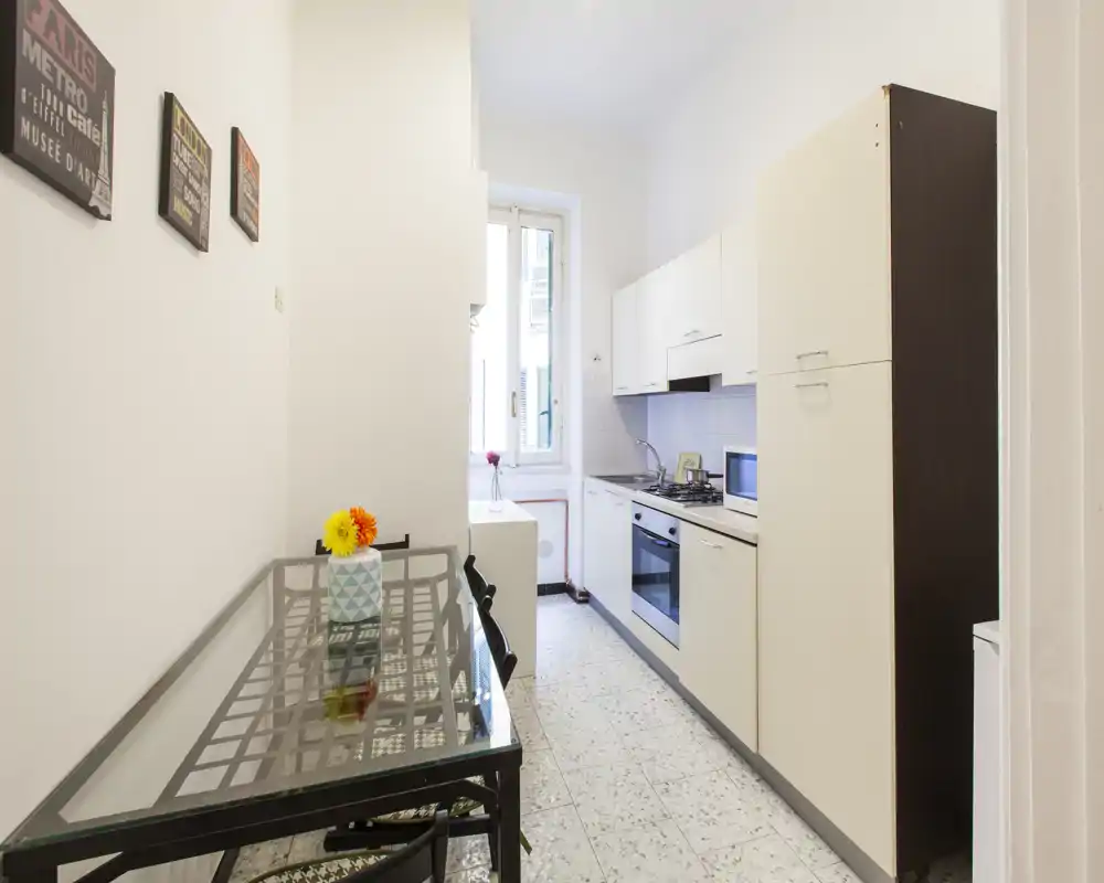 Cucina stretta e luminosa con pensili bianchi, forno integrato e tavolo con piano in vetro; finestra porta luce naturale e un piccolo vaso decorativo.