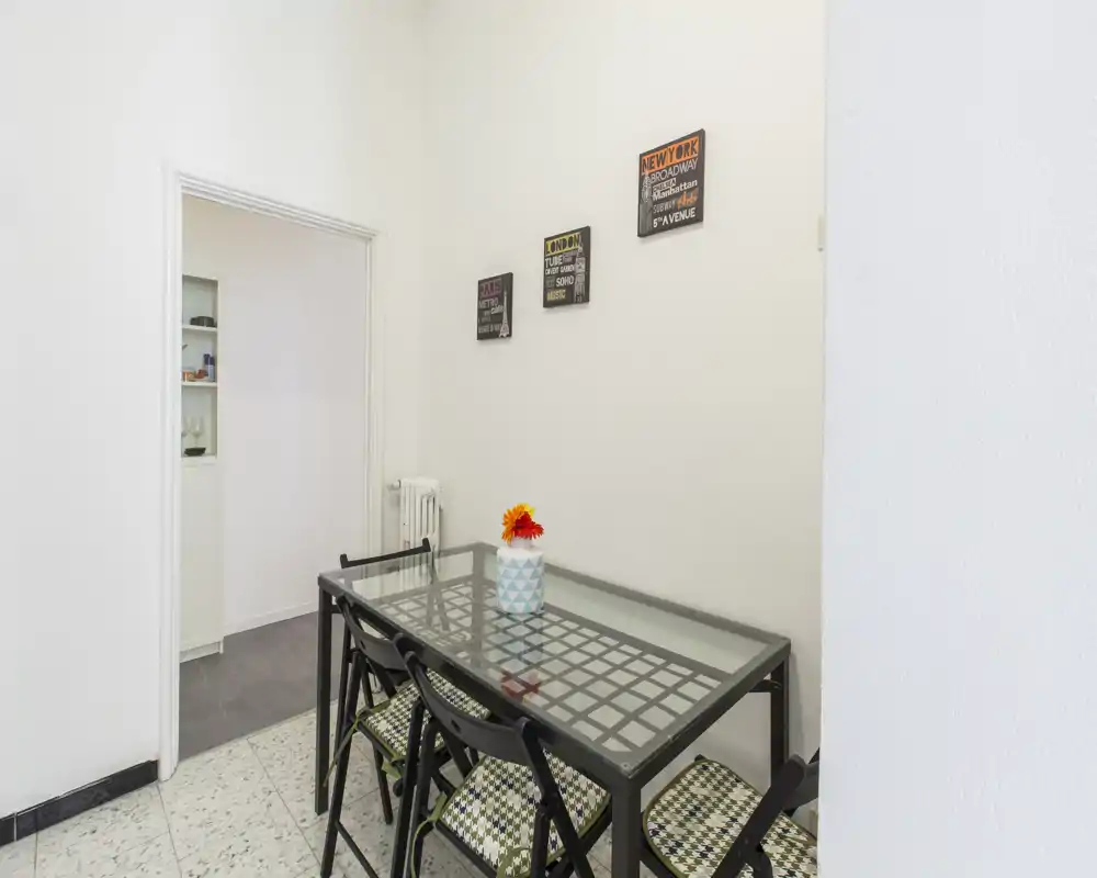 Zona pranzo compatta con tavolo con piano in vetro, quattro sedie pieghevoli con cuscini a fantasia e un vaso decorativo; luminosa, ordinata e adatta a un piccolo appartamento.