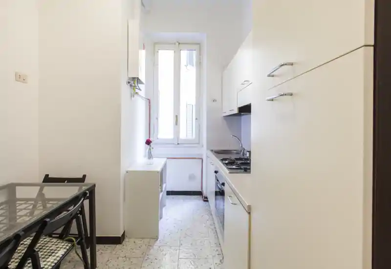 Compact galley kitchen with white cabinets, gas hob, built-in oven and a window providing natural light. Small dining table and simple decor create an efficient, bright space.