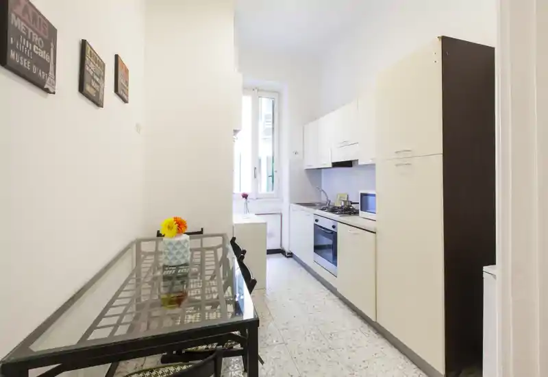 Bright narrow kitchen with white cabinets, built-in oven and a glass-topped dining table; small window provides natural light and a modest decorative vase.