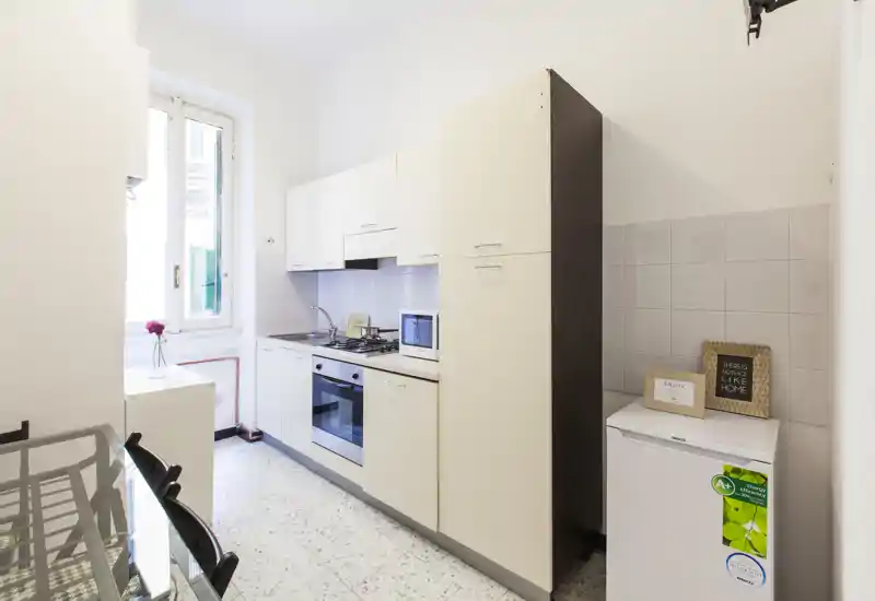 Compact, bright fitted kitchen with white cabinets, built-in oven and gas hob, small fridge and a window providing natural light.