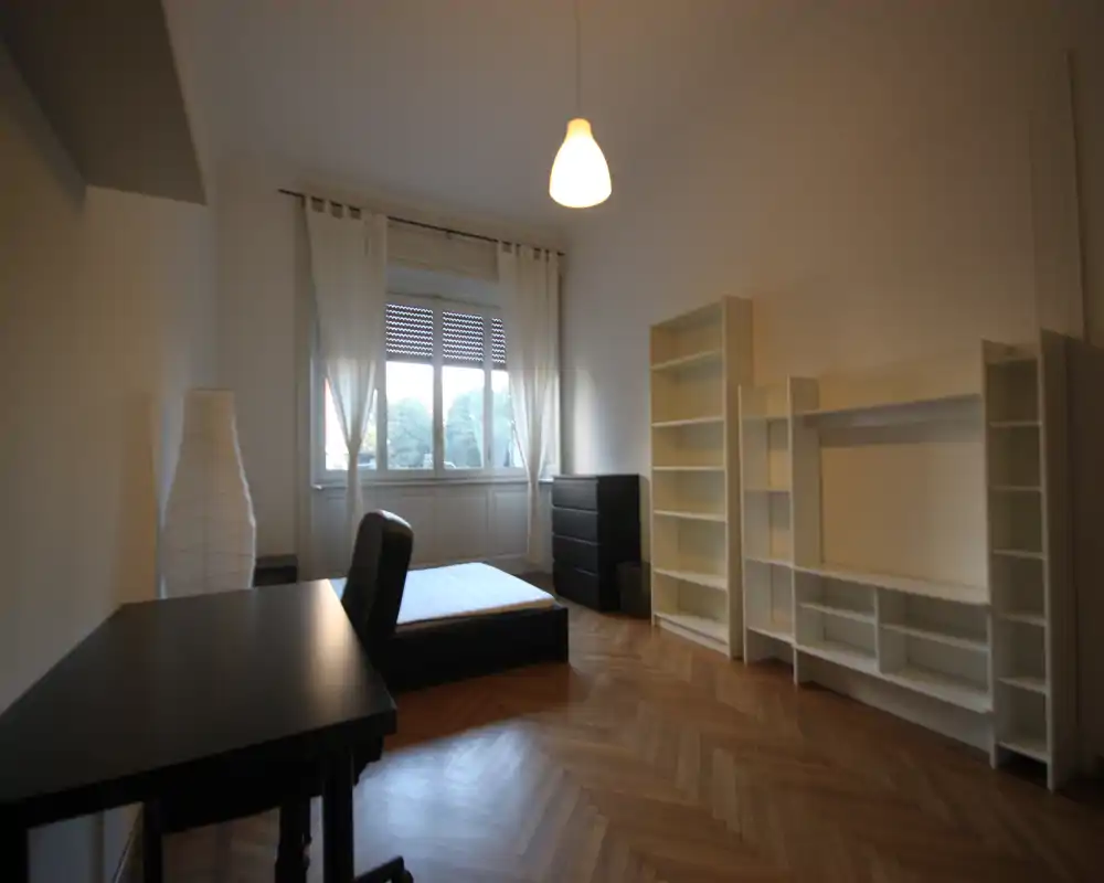 Dormitorio minimalista y luminoso con suelo de parquet, amplia ventana y estanterías; incluye cama baja y escritorio, ideal para un alquiler moderno.