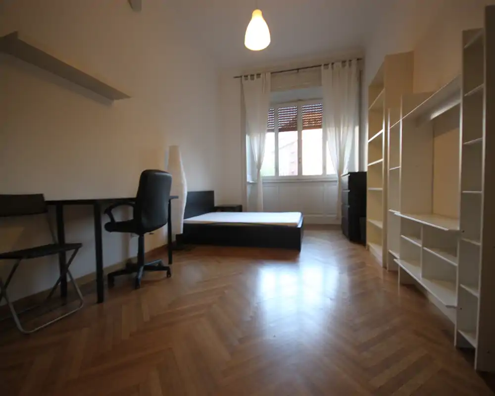 Dormitorio luminoso y minimalista con cama baja doble, escritorio y estanterías; suelo de parquet y gran ventana que aporta luz natural.
