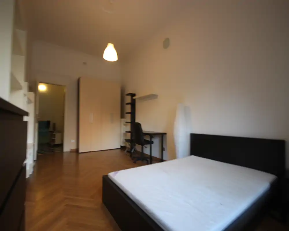 Amplio dormitorio con cama doble en primer plano, suelo de parquet, armario y pequeño escritorio. Luminoso y amueblado de forma sencilla, apto para una estancia cómoda.