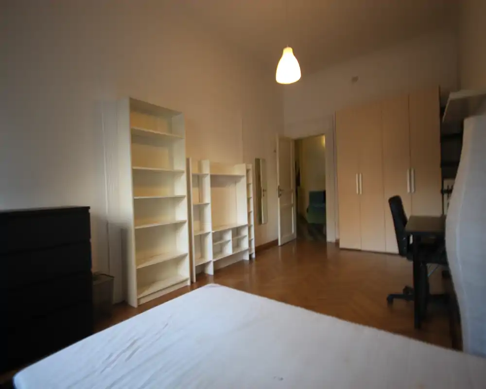 Dormitorio amplio y luminoso con colchón visible en primer plano, estanterías altas, armario y escritorio; lámpara de techo cálida y suelo de parquet crean un espacio acogedor y ordenado.