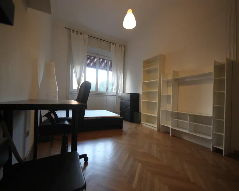 Dormitorio luminoso y minimalista con cama baja junto a una gran ventana, escritorio y estanterías; suelo de parquet y cortinas neutras aportan sensación de amplitud.