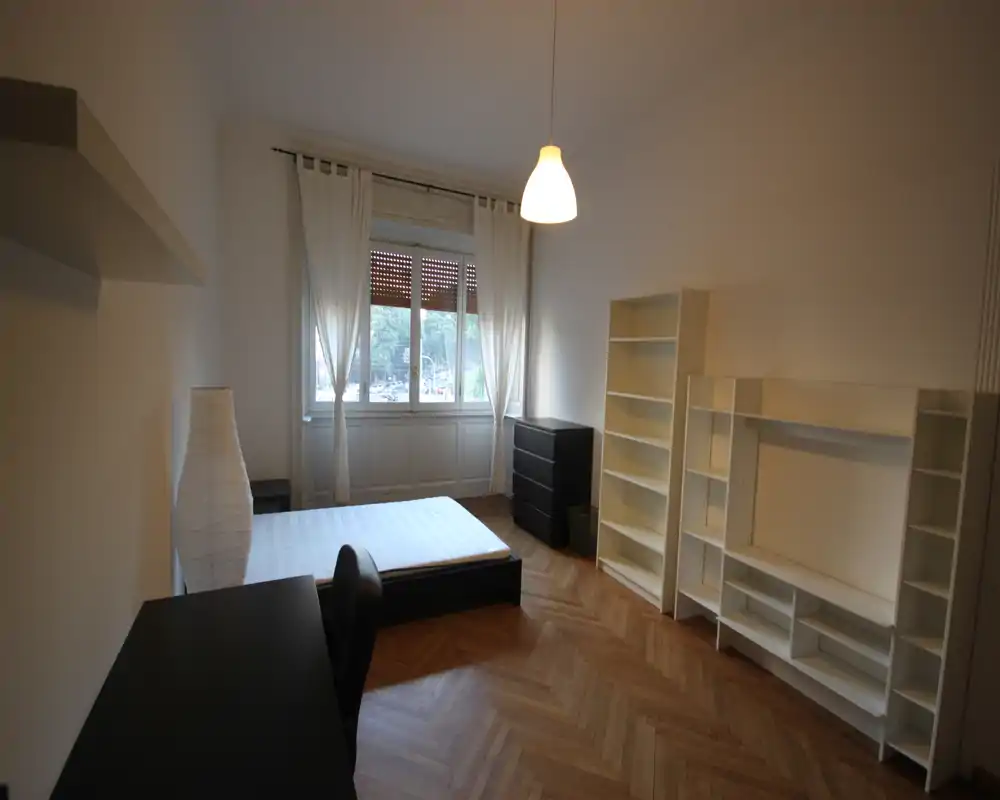 Dormitorio luminoso y sencillamente amueblado con cama doble, escritorio y estanterías; gran ventana con luz natural y suelo de parquet en espiga.