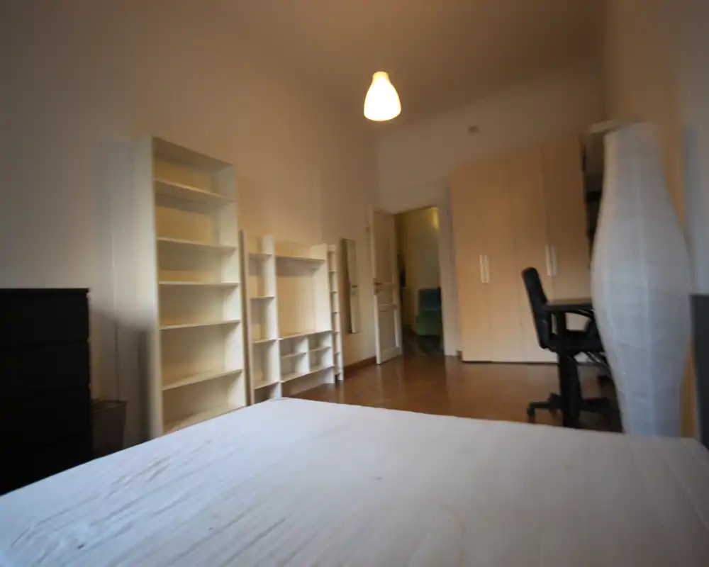 Dormitorio luminoso y de mobiliario sencillo con colchón en primer plano, estanterías y armario en la pared, y un escritorio con silla a la derecha. Tonos neutros y suelo de parquet que transmiten calma.
