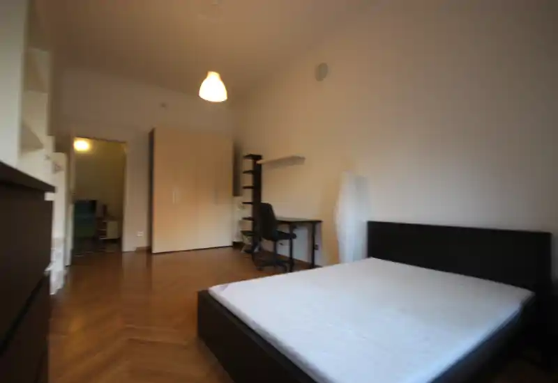 Ampia camera da letto con letto matrimoniale in primo piano, pavimento in parquet, armadio e una scrivania. Luminosa e arredata in modo essenziale, adatta a un soggiorno confortevole.