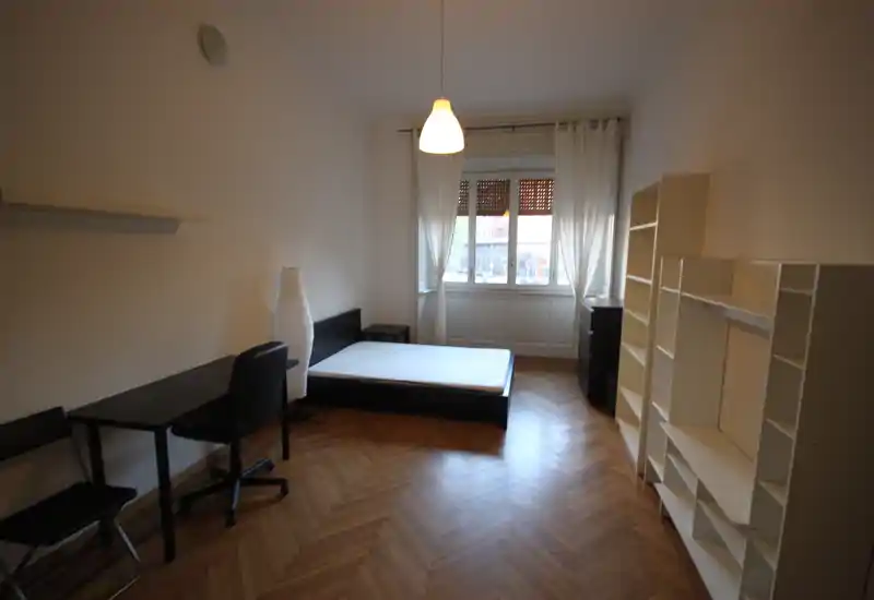 Dormitorio luminoso y sencillo con cama doble sobre estructura baja, escritorio y estanterías; amplia ventana que aporta luz natural al suelo de parquet.
