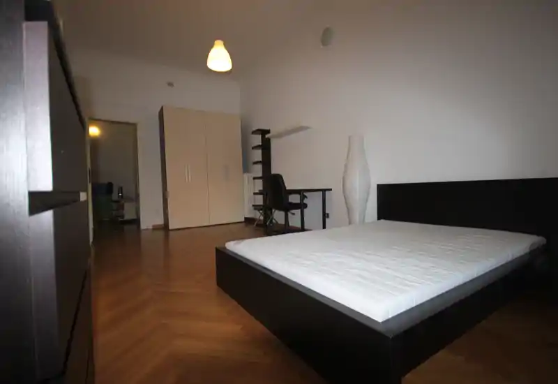 Dormitorio luminoso y minimalista con cama tipo plataforma oscura, suelo de parquet, armario y pequeño escritorio: líneas modernas y buena luz natural.