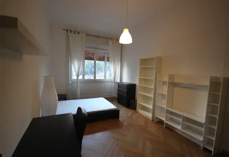 Camera da letto luminosa e arredata in modo essenziale con letto matrimoniale, scrivania e scaffalature; grande finestra e pavimento in parquet a spina di pesce.