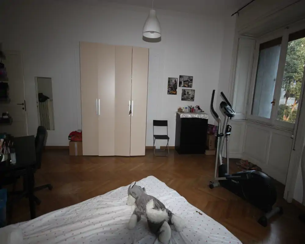 Dormitorio amplio con la cama en primer plano, suelo de parqué, armario contra la pared y bicicleta estática junto a la ventana. La gran ventana aporta luz natural; la estancia se siente luminosa y despejada.