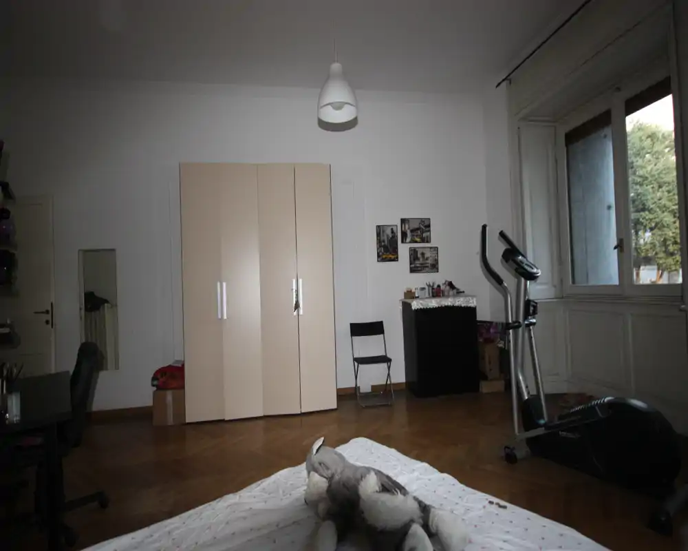 Dormitorio amplio con el colchón visible en primer plano, suelos de parquet y una gran ventana que aporta luz natural. Armario de tonos neutros y mobiliario sencillo que crean un espacio funcional y tranquilo.