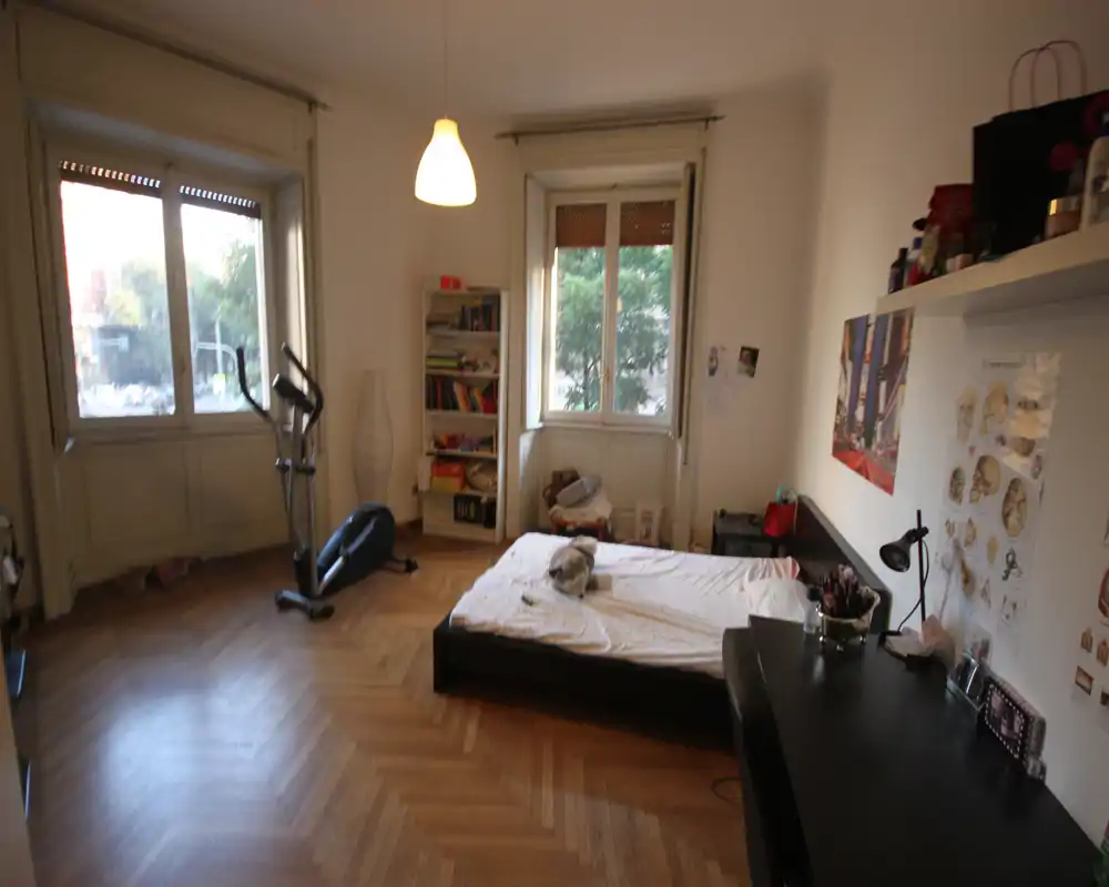 Habitación amplia con cama baja, grandes ventanas que permiten abundante luz natural, zona de escritorio y estanterías. Suelo de parqué y decoración personal mínima crean un ambiente acogedor y vivido.