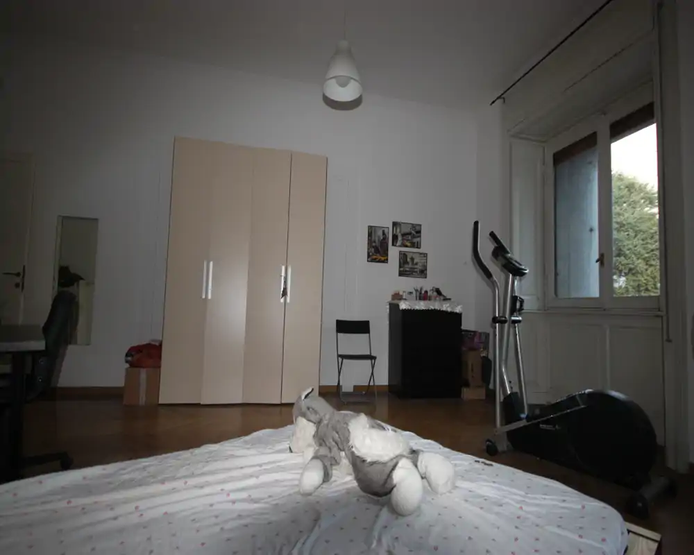 Dormitorio luminoso y poco amueblado con la cama en primer plano, armario, cómoda y una bicicleta estática junto a la ventana. La ropa de cama y un peluche aportan un toque acogedor y la ventana ofrece luz natural.