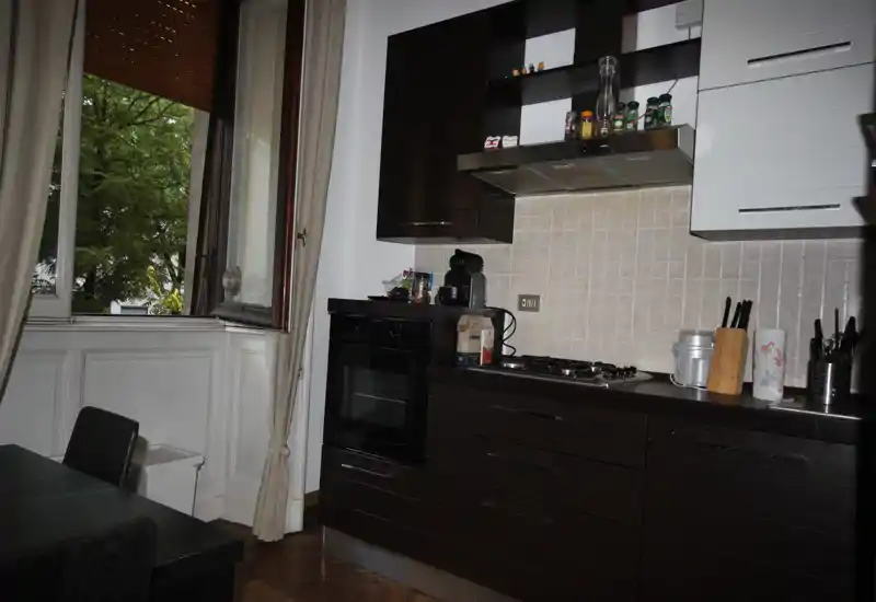 Angolo cucina moderno con mobili in legno scuro, forno incassato e piano cottura a gas, piccoli elettrodomestici sul piano e una grande finestra aperta che illumina naturalmente.