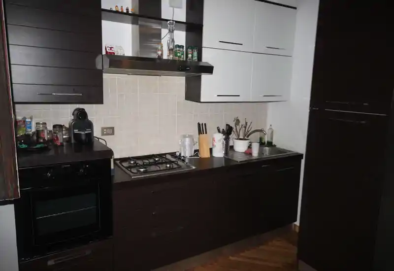 Cucina moderna compatta con pensili bianchi e basi scure, piano cottura a gas, forno incassato e lavello; ripiani ordinati con piccoli elettrodomestici e pianta.