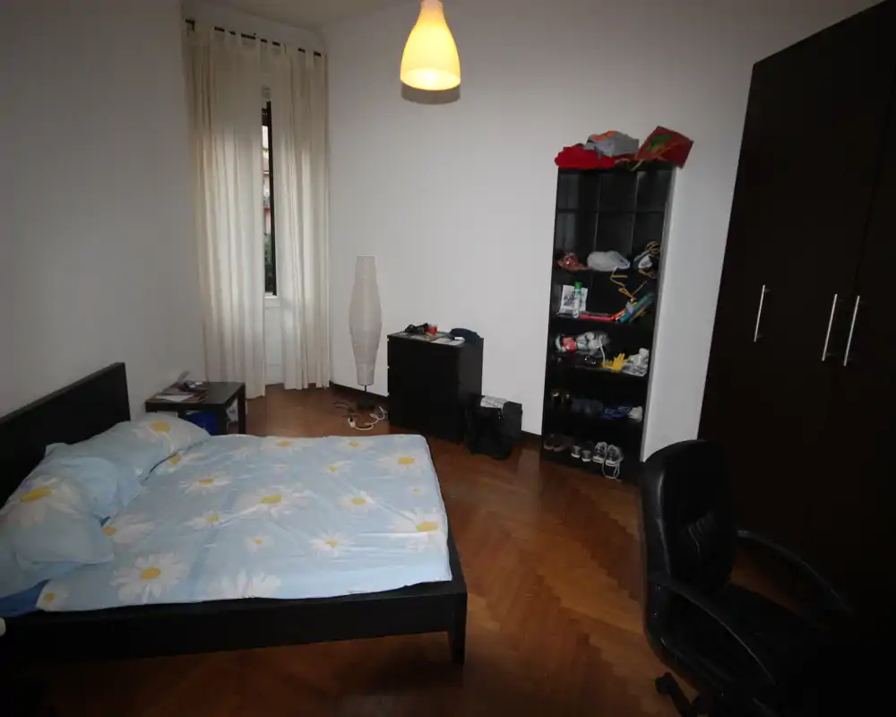 Simple furnished bedroom with a double bed on a dark frame, parquet flooring, wardrobe and shelving; soft pendant light and a window with curtains provide natural and warm lighting.