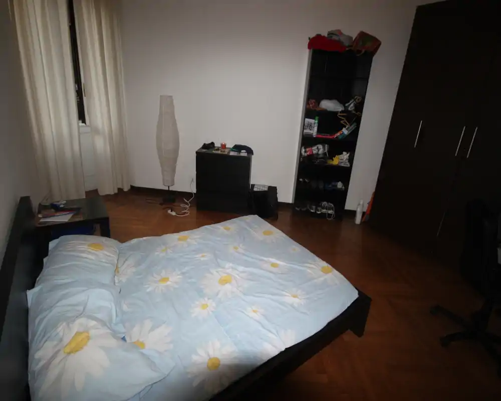 Bright double bedroom with a floral duvet, wooden parquet floor, wardrobe and shelving. The room is simply furnished and well-lit by a window with light curtains.
