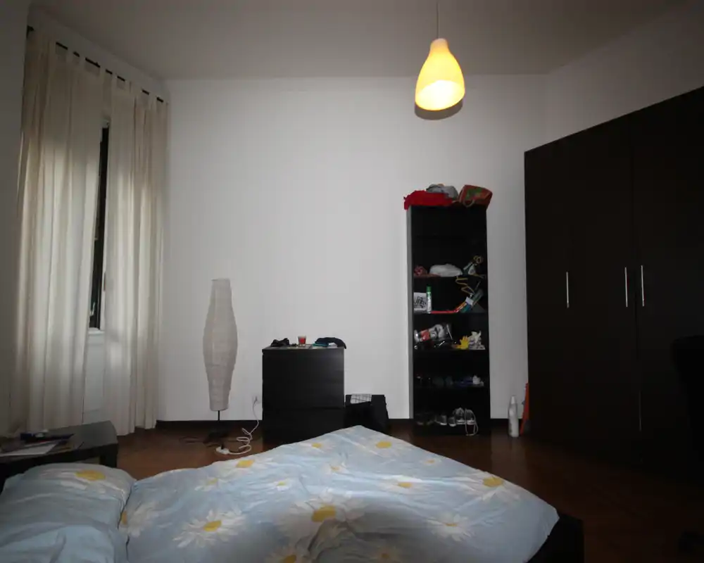 Bright, simply furnished bedroom with a double bed in the foreground, a tall wardrobe and shelving unit against the far wall, and soft overhead lighting.