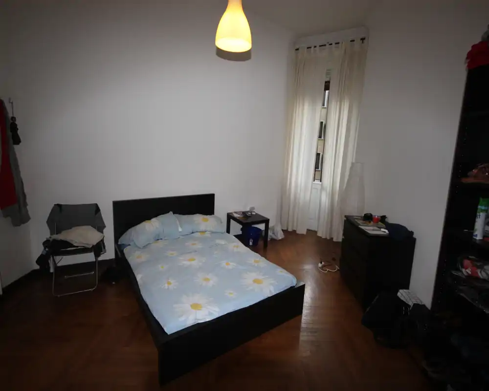 Simple, well-lit bedroom with a double bed on a dark frame, wooden parquet floor and a tall window with light curtains. Functional furnishings and soft bedding create a comfortable, uncluttered space ideal for rental listings.