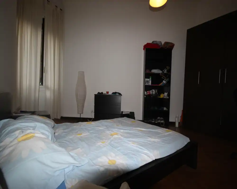 Bright, simply furnished bedroom with a large double bed in the foreground, a tall lamp, shelving unit and a wardrobe along the wall.