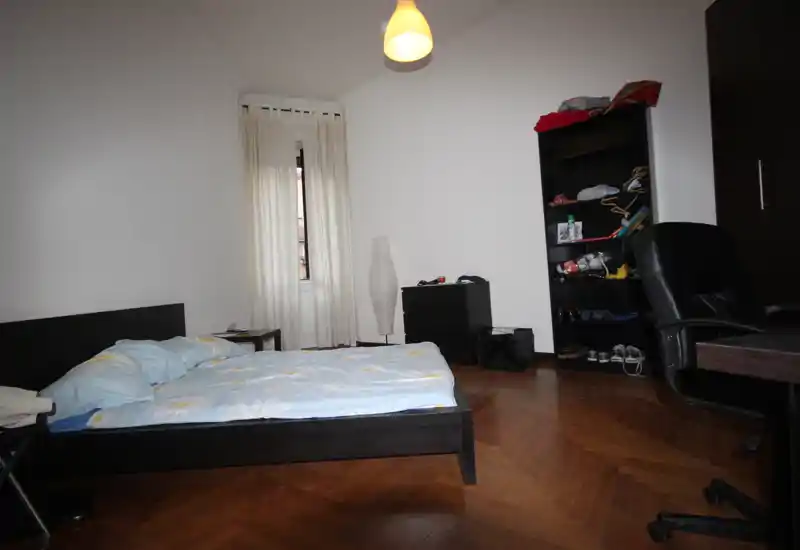Chambre claire et simple avec un grand lit bas, parquet, fenêtre avec voilages et mobilier minimal comprenant une étagère et une chaise de bureau.