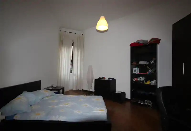 Chambre simple et lumineuse avec un lit double, table de chevet, lampadaire et étagère. La lumière venant de la fenêtre et les murs blancs offrent une atmosphère calme, idéale pour une location.