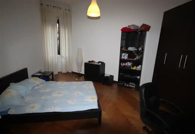 Dormitorio sencillo y bien iluminado con cama doble en estructura de madera oscura, suelo de parquet y un amplio armario. Incluye mesita, estantería y lámpara colgante que aportan un espacio funcional.