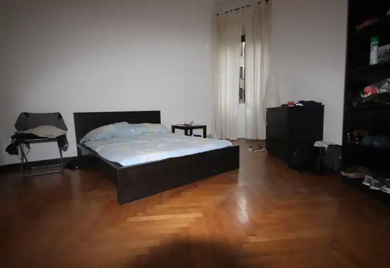 Chambre spacieuse avec lit double bas sur parquet en chevrons, table de chevet simple et lumière naturelle provenant d'une haute fenêtre.