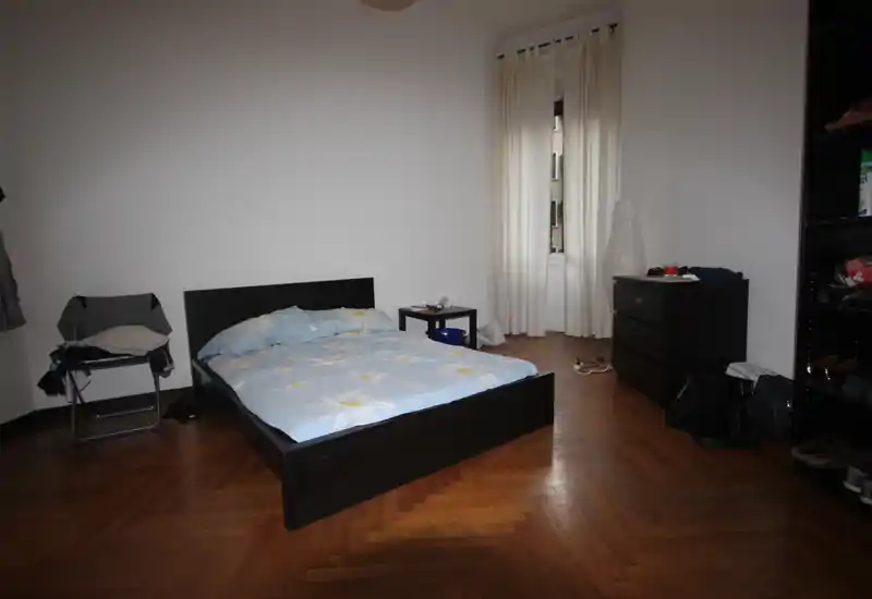 Chambre spacieuse avec lit double, parquet et une fenêtre munie de rideaux clairs. Agencement sobre avec une commode et des étagères pour le rangement.
