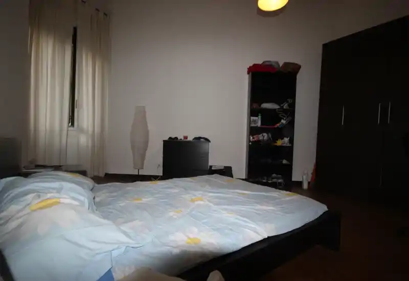 Dormitorio luminoso y sencillo con cama doble con ropa de cama azul con flores, lámpara de pie, estantería y gran armario.