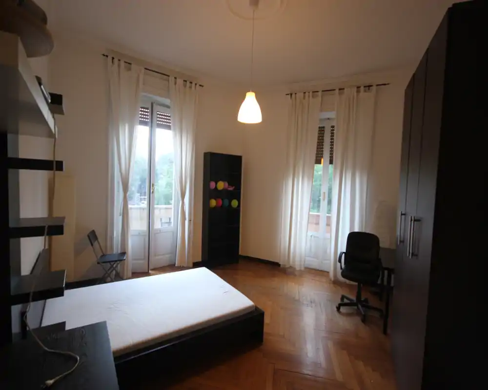 Spacious, bright bedroom with a low-profile double bed, two tall French windows with light curtains, parquet flooring and a large wardrobe. Simple modern furnishings and good natural light make it suitable for rental listings.