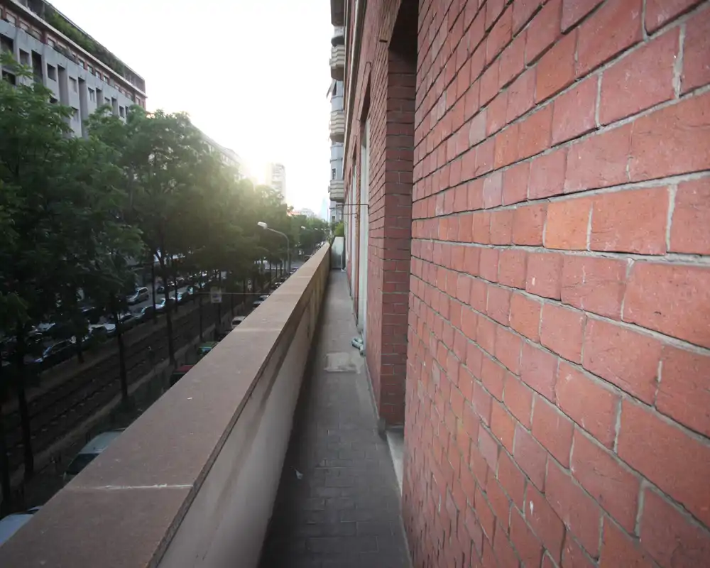 Narrow balcony running along the building facade with brick wall on one side and a low railing overlooking a tree-lined street. Good for a quick step outside and city views.