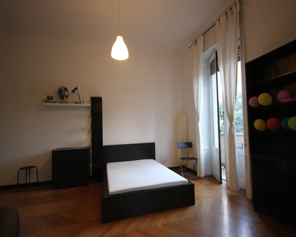 Spacious bedroom with a low-profile double bed, large window with sheer curtains and warm parquet flooring. Simple, modern shelving and soft natural light make the room airy and functional for rest.