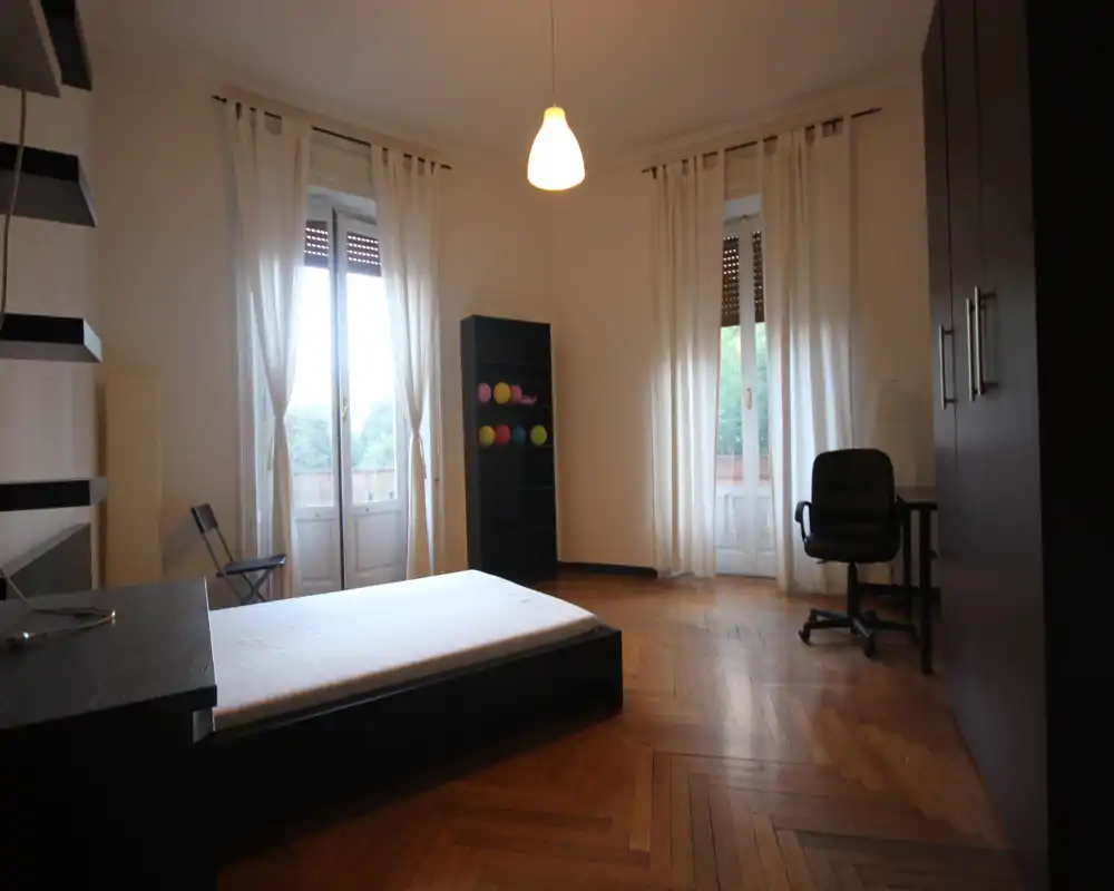 Spacious bedroom with parquet flooring, low platform bed near two tall windows letting in natural light, wardrobe and small desk area — bright, airy and simply furnished.