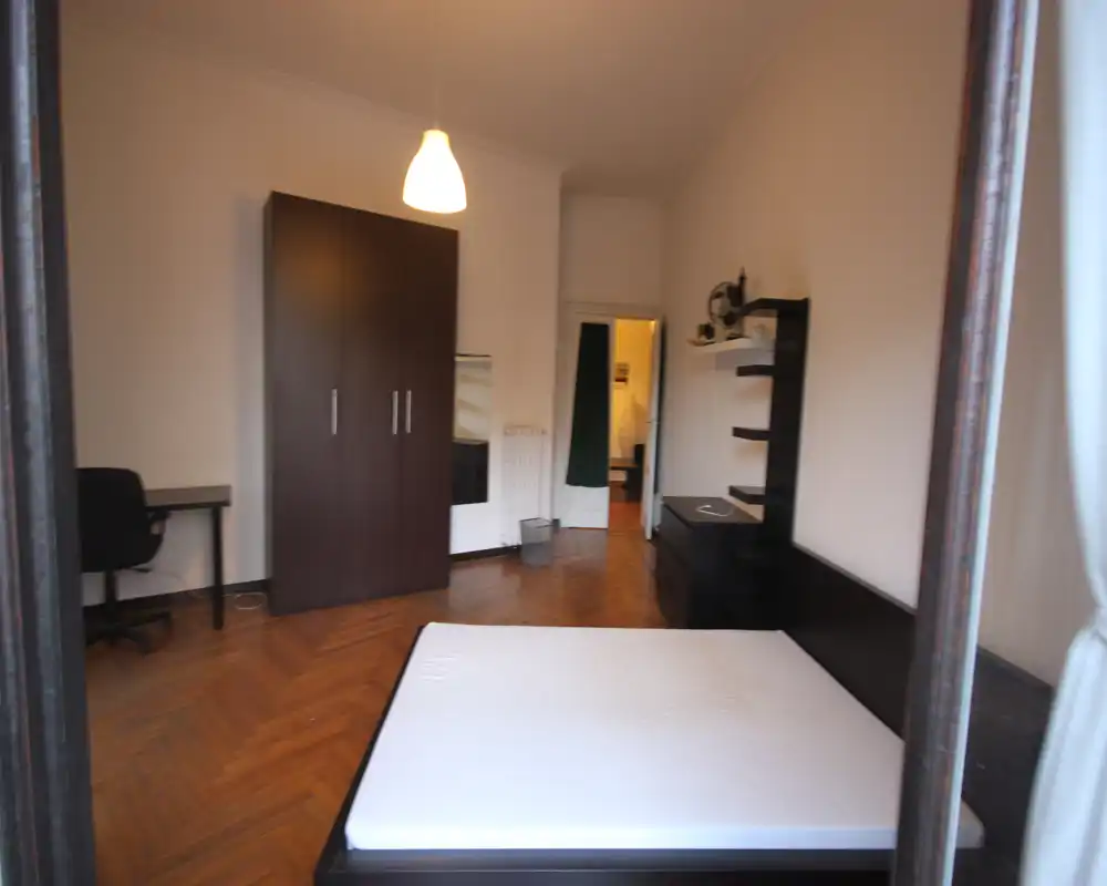 Bright, well-arranged bedroom with a double bed in the foreground, wardrobe, desk and shelving; hardwood parquet floor and natural light from the window.