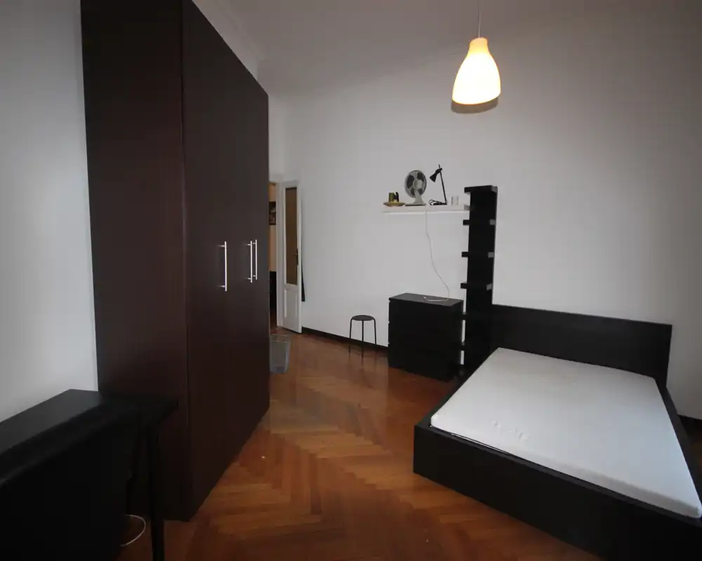 Bright, minimally furnished bedroom with a visible double bed, dark wood furniture and herringbone parquet flooring. Neutral white walls and a pendant light create a clean, modern look suitable for rental photos.