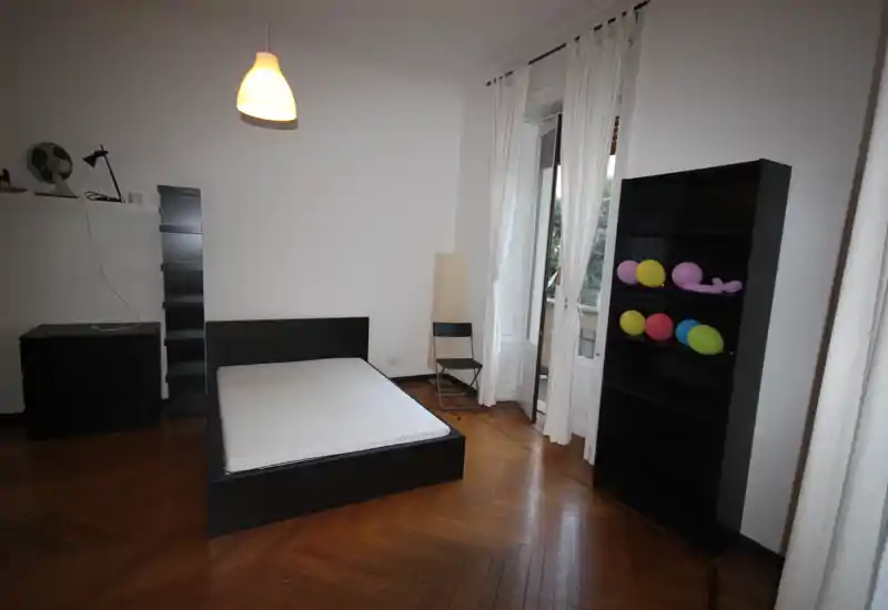 Spacious bedroom with a low platform double bed, large window with white curtains letting in natural light, parquet flooring and minimalist black furniture.
