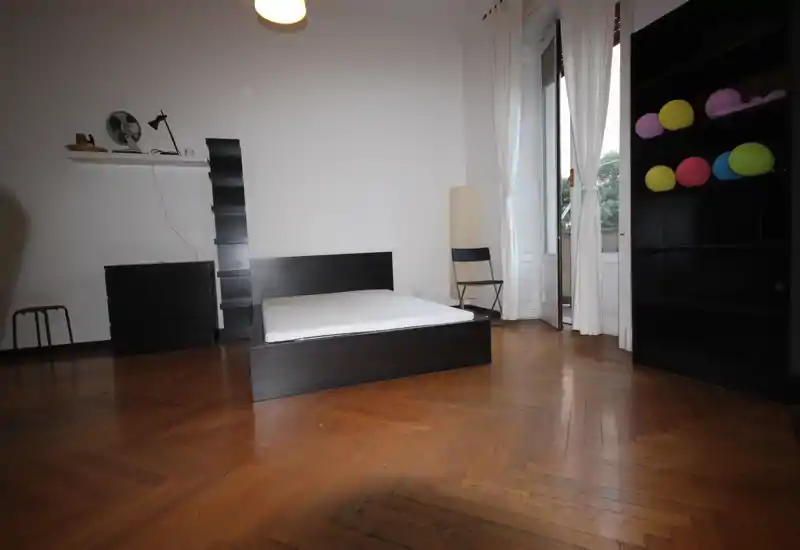 Spacious bedroom with a low-profile double bed, tall windows draped in light curtains, parquet floors and minimalist dark furniture. Natural light fills the room creating an airy, modern feel.