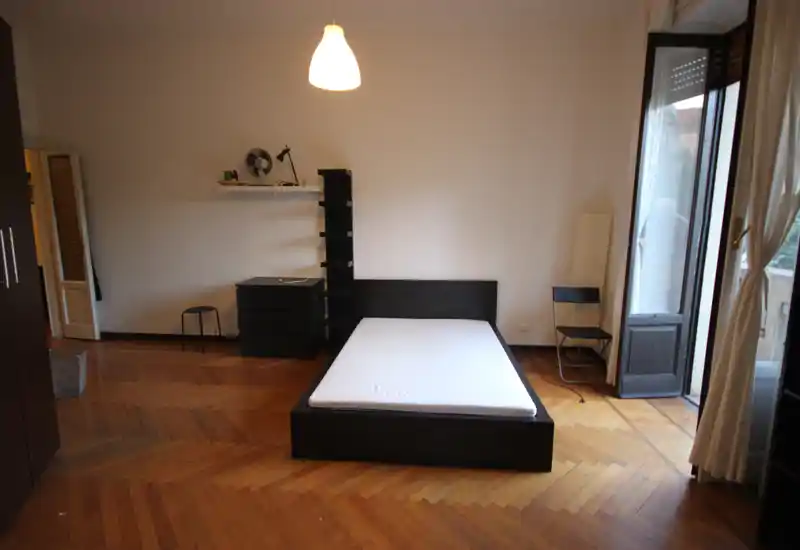 Bright, minimal bedroom with a central double bed on a low dark frame, parquet floor and a balcony door letting in natural light.