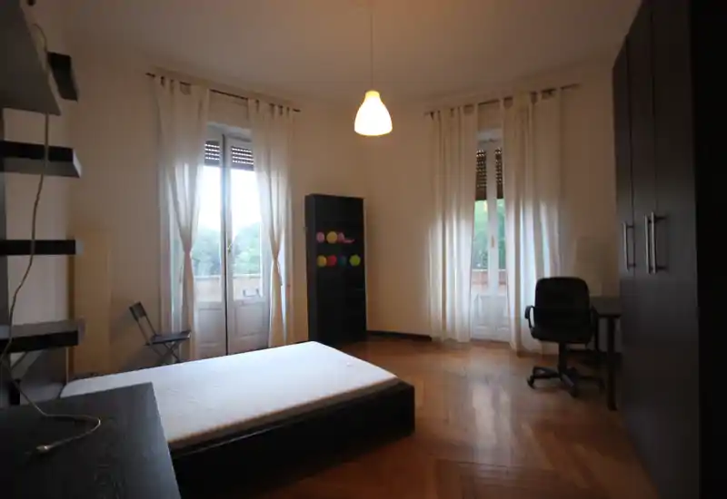 Spacious bedroom with high ceilings, two tall windows and a simple low bed frame. Natural light filters through sheer curtains, complemented by wooden parquet floors and minimal, modern furniture.