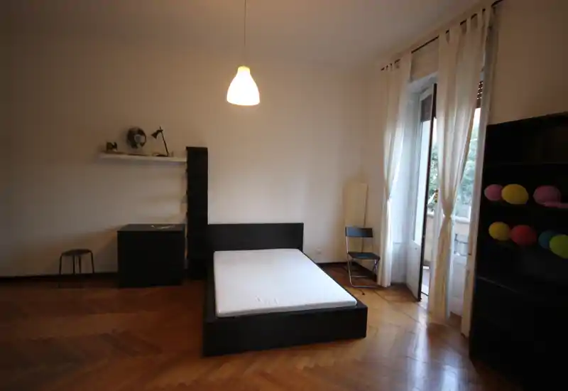 Spacious bedroom with a double bed on a dark wood frame, parquet floors and a balcony allowing natural light. Minimalist furnishings include shelving, a chest of drawers and a chair, creating a calm, airy atmosphere.